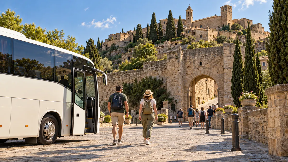 A white coach parked on a cobbled forecourt while travellers walk towards the arched gateway of a walled hilltop town.
